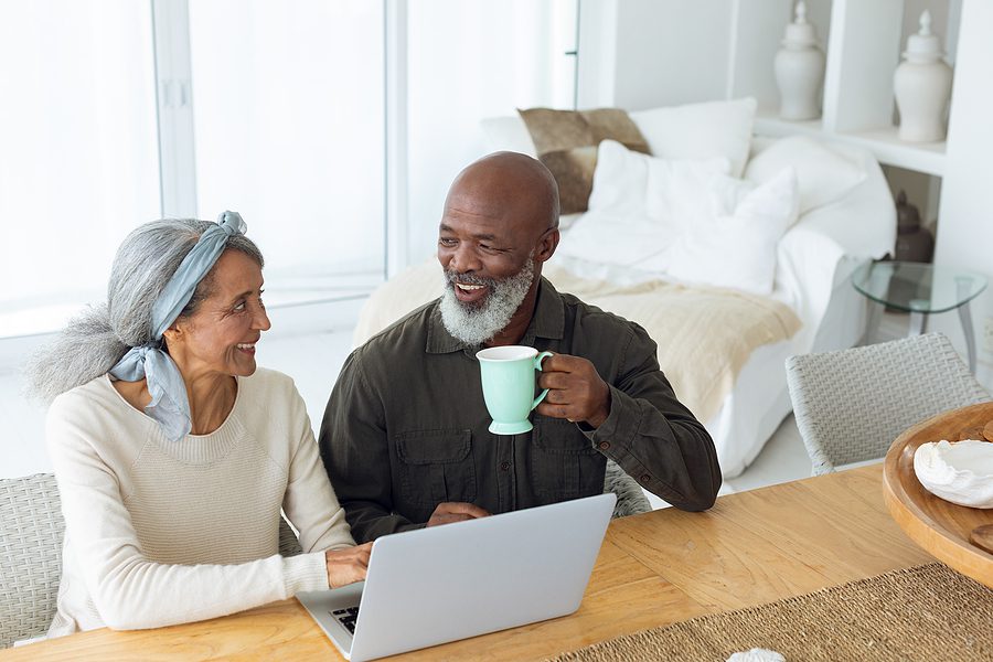 Understanding Cost of Senior Independent Living Front view of diverse senior couple using laptop on table while man holds a cup in beach house.