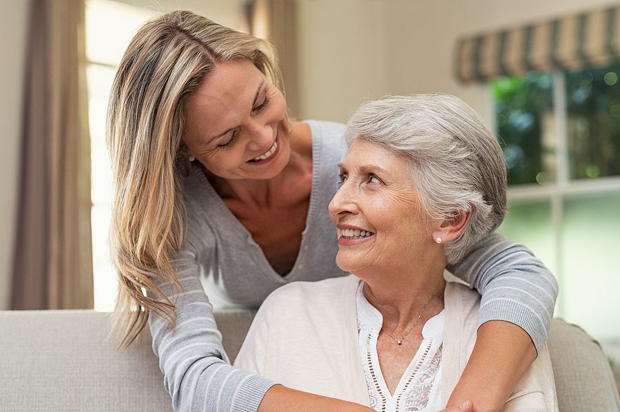 Adult daughter hugs older adult mother on couch in living room Adult daughter and older adult mother hug after discussing how to maintain healthy family relationships
