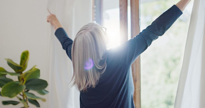 Older adult woman pulling back blinds to let morning sunshine in